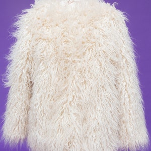 May include: A white faux fur jacket with a long, shaggy pile. The jacket is on a mannequin and has a relaxed fit.
