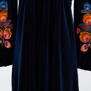 May include: A dark blue velvet dress with long sleeves and colourful floral embroidery on the sleeves. The dress features a round neckline with blue embroidery and tassels. The fabric has a soft, flowing drape.
