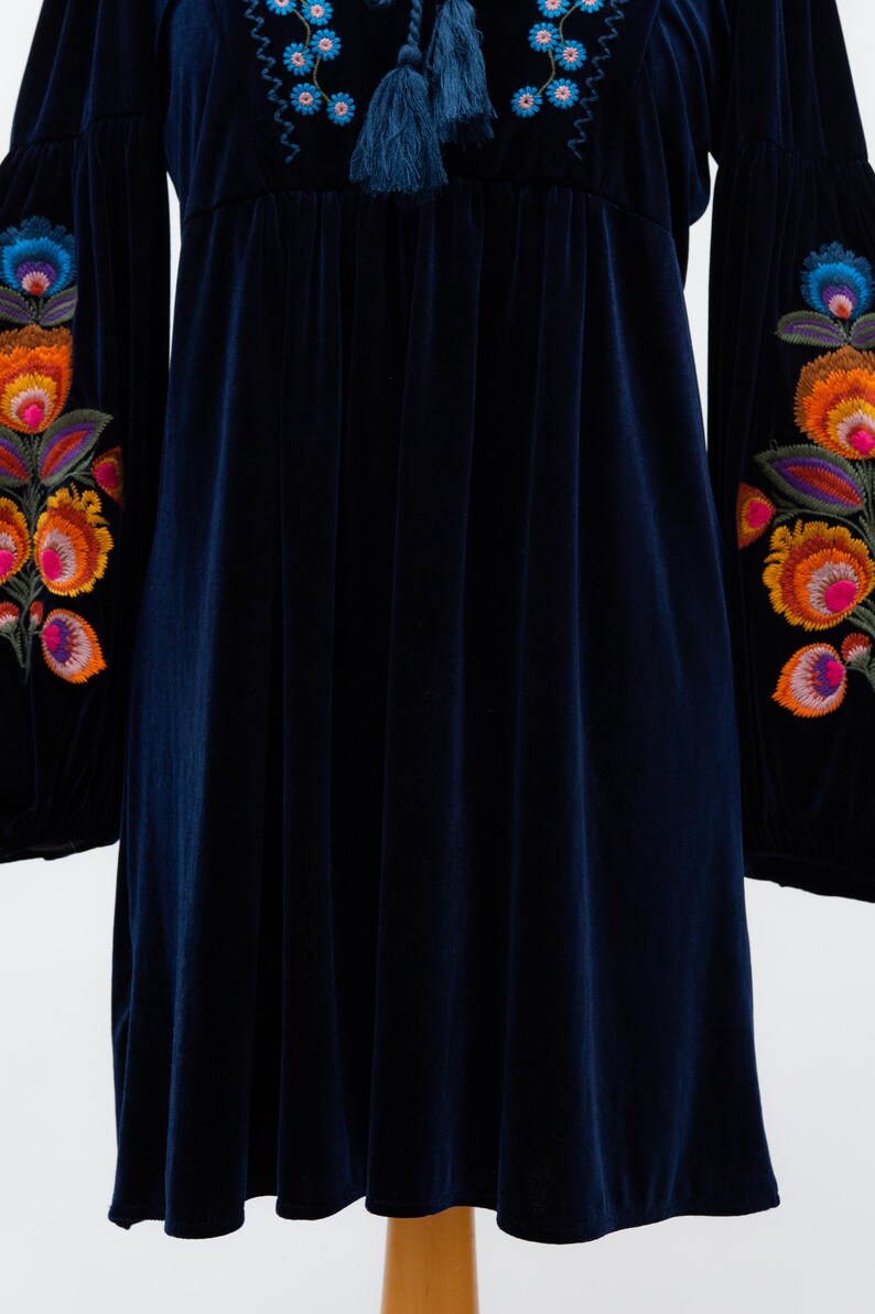 May include: A dark blue velvet dress with long sleeves and colourful floral embroidery on the sleeves. The dress features a round neckline with blue embroidery and tassels. The fabric has a soft, flowing drape.
