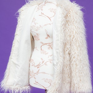 May include: A white faux fur jacket with long sleeves and a fluffy texture. The jacket is open and draped over a white mannequin with a floral pattern.