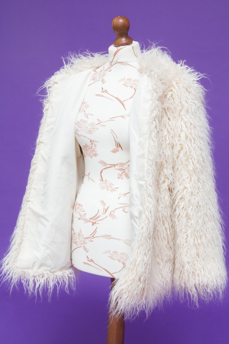 May include: A white faux fur jacket with long sleeves and a fluffy texture. The jacket is open and draped over a white mannequin with a floral pattern.