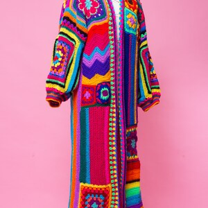 K&ouml;nnte beinhalten: Ein farbenfroher, langer H&auml;kelmantel auf einer Schaufensterpuppe. Der Mantel hat ein Patchwork-Design mit Rosa, Rot, Gelb, Blau und Gr&uuml;n. Die &Auml;rmel sind lang und puffig, und der Mantel hat ein Blumenmuster auf den Schultern. Ma&szlig;e sind nicht verf&uuml;gbar.