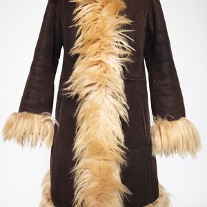 May include: A long, dark brown coat with a faux fur collar, cuffs, and trim. The coat has a vintage style with a hood and a tailored fit. The faux fur is a light tan color, creating a striking contrast with the dark brown fabric.