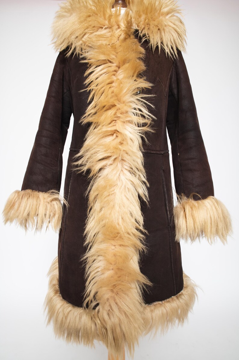 May include: A long, dark brown coat with a faux fur collar, cuffs, and trim. The coat has a vintage style with a hood and a tailored fit. The faux fur is a light tan color, creating a striking contrast with the dark brown fabric.