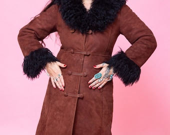 May include: A long brown suede coat with a black sheepskin collar and cuffs. The coat has a button closure and a fitted silhouette.