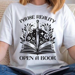 May include: White t-shirt with a black and white graphic design. The design features an open book with a sword, flowers, and the text "PAUSE REALITY OPEN A BOOK". The shirt is made of a soft material.