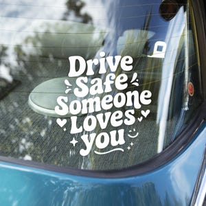 May include: White vinyl decal with the text "Drive Safe Someone Loves You" in a whimsical font, adorned with hearts, stars, and a smiling face. The decal is applied to a car window, with a blurred background.