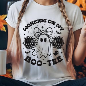May include: White t-shirt with a black graphic of a ghost lifting weights, a bow with a skull, and the text "WORKING ON MY BOO-TEE". The design includes bats and stars, perfect for Halloween or fitness enthusiasts.