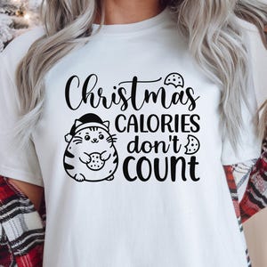 May include: White t-shirt with the text "Christmas calories don't count." A cartoon cat wearing a Santa hat and holding a cookie is featured in the design. The shirt also includes SVG, PNG, EPS, and DXF file format icons.
