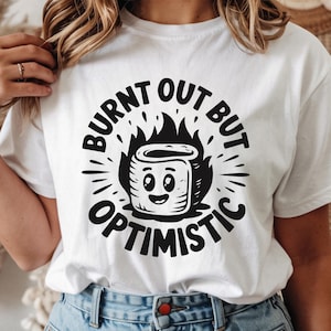 Puede incluir: Camiseta blanca con un diseño gráfico negro que presenta una malvavisco sonriente en llamas, con las palabras "BURNT OUT BUT OPTIMISTIC" en un diseño circular. La camiseta también tiene iconos de tipo de archivo SVG, PNG, EPS y DXF.