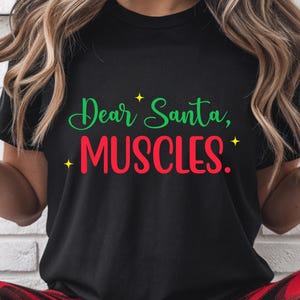 May include: Black t-shirt with the text "Dear Santa, MUSCLES." in green and red, with yellow star accents. The shirt is a casual style.