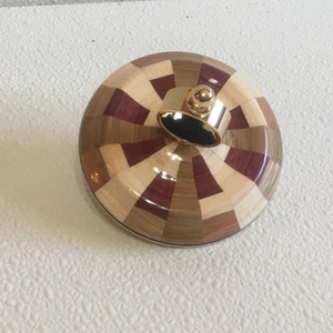 May include: A wooden spinning top with a geometric pattern of light and dark wood. The top has a gold metal ring with a small ball on top.