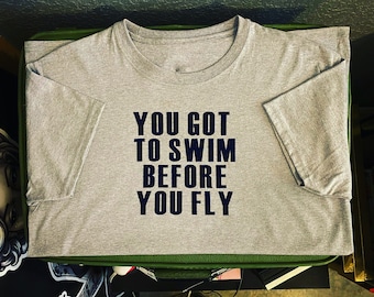 Op de afbeelding: Een grijs T-shirt met de tekst "YOU GOT TO SWIM BEFORE YOU FLY" in zwart gedrukt.