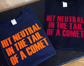 Op de afbeelding: Twee marineblauwe t-shirts met de tekst "HIT NEUTRAL IN THE TAIL OF A COMET" in oranje en rood. De tekst is vetgedrukt en gecentreerd op de shirts.