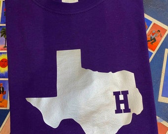 Op de afbeelding: Paars T-shirt met een witte omtrek van de staat Texas en een witte letter "H" binnen de omtrek.
