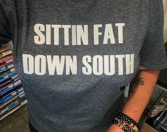 Op de afbeelding: Een grijs T-shirt met de tekst "Sittin Fat Down South" in wit gedrukt. De persoon die het shirt draagt, draagt ook een parelketting en meerdere armbanden.
