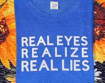 Op de afbeelding: Een koninklijk blauw t-shirt met witte tekst die "REAL EYES REALIZE REAL LIES" luidt.