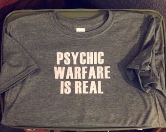 Op de afbeelding: Een donkergroen T-shirt met de tekst "PSYCHIC WARFARE IS REAL" in wit gedrukt op de voorkant.