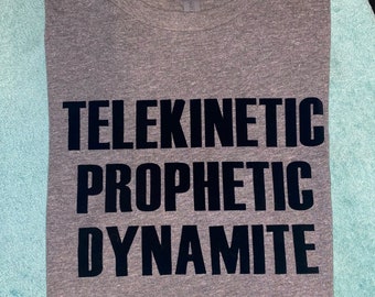 Op de afbeelding: Een heather grijze t-shirt met de woorden "TELEKINETIC PROPHETIC DYNAMITE" in vette zwarte letters. Het shirt is netjes opgevouwen, waardoor de tekst opvalt. De stof lijkt zacht en comfortabel.