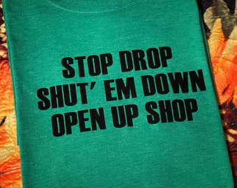 Op de afbeelding: Groen T-shirt met zwarte tekst die "Stop Drop Shut 'Em Down Open Up Shop" luidt.