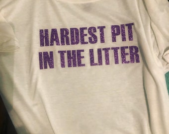 Op de afbeelding: Wit T-shirt met paarse glittertekst die "Hardest Pit in the Litter" zegt.