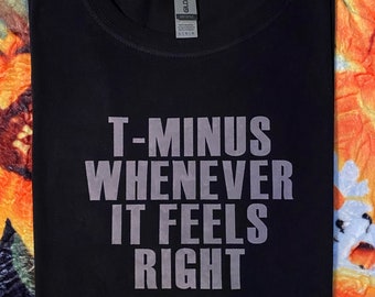 Op de afbeelding: Zwart T-shirt met de tekst "T-Minus Whenever It Feels Right" in lichtgrijs gedrukt.
