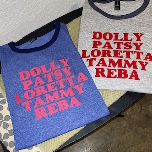 May include: Two blue and gray t-shirts with a white ringer. The shirts have the names of famous female country singers printed in red: Dolly, Patsy, Loretta, Tammy, and Reba.