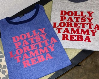 Op de afbeelding: Twee blauwe en grijze T-shirts met een witte boord. De shirts hebben de namen van beroemde vrouwelijke countryzangeressen in rood gedrukt: Dolly, Patsy, Loretta, Tammy en Reba.