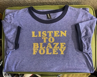 Op de afbeelding: Een blauw t-shirt met een donkerblauwe boord en gouden glitterletters met de tekst "Listen to Blaze Foley".
