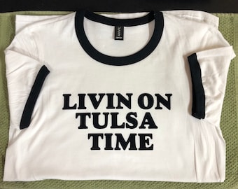 Op de afbeelding: Wit T-shirt met zwarte ringerrand en de tekst "Livin on Tulsa Time" in zwart gedrukt.