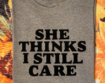 Op de afbeelding: Grijs T-shirt met zwarte tekst die "Ze denkt dat ik nog steeds om haar geef." zegt.