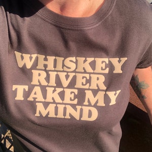 Peut inclure: Un t-shirt gris fonc&eacute; avec le texte "Whiskey River Take My Mind" imprim&eacute; en beige clair.