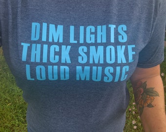 Op de afbeelding: Een donkerblauw t-shirt met de tekst "DIM LIGHTS THICK SMOKE LOUD MUSIC" in lichtblauw. Het t-shirt wordt gecombineerd met een blauwe jeans. Een bloementattoo is zichtbaar op de arm van de persoon.