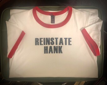 Op de afbeelding: Wit ringer-t-shirt met rode randen op de kraag en mouwen. De tekst "REINSTATE HANK" is in zwarte blokletters op de voorkant van het shirt gedrukt. Het shirt ligt op een groene ondergrond.