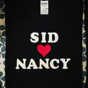 May include: Black t-shirt with white text that reads "SID" with a red heart and "NANCY" underneath.