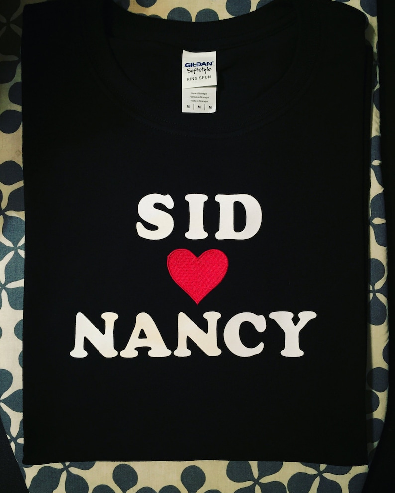 May include: Black t-shirt with white text that reads "SID" with a red heart and "NANCY" underneath.