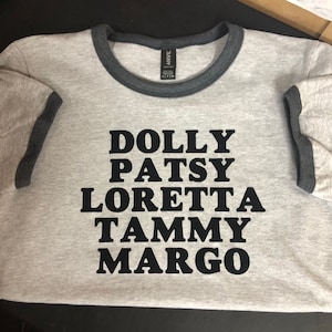 May include: A gray t-shirt with a black ringer and the names "DOLLY PATSY LORETTA TAMMY MARGO" printed in black on the front.