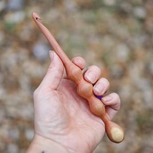 May include: A handcrafted wooden crochet hook with a light brown finish. The hook features a curved end and a bulbous handle with a series of rounded shapes. The hook is held in a hand, showcasing its ergonomic design.