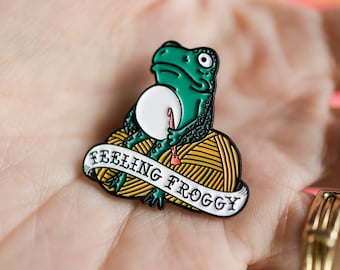 Op de afbeelding: Groene emaille pin met een kikker die een rode breinaald en een witte bol wol vasthoudt. De kikker zit op een bol gele wol met een wit lint waarop "Feeling Froggy" staat.