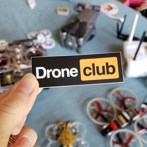 May include: A black and yellow sticker with the text "Drone club" on it. The sticker is being held by a hand.