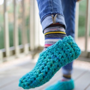 May include: A pair of teal blue crocheted slippers with a textured knit pattern. The slippers are worn on a person's feet with jeans and striped socks.
