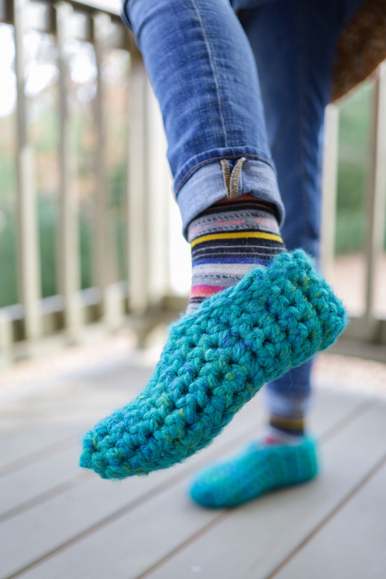 May include: A pair of teal blue crocheted slippers with a textured knit pattern. The slippers are worn on a person's feet with jeans and striped socks.
