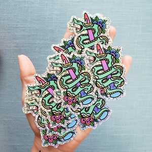 May include: A handful of holographic stickers featuring a green snake with a pink dagger through its body, surrounded by flowers and leaves. The stickers have a glittery, iridescent finish and are on a light blue background.