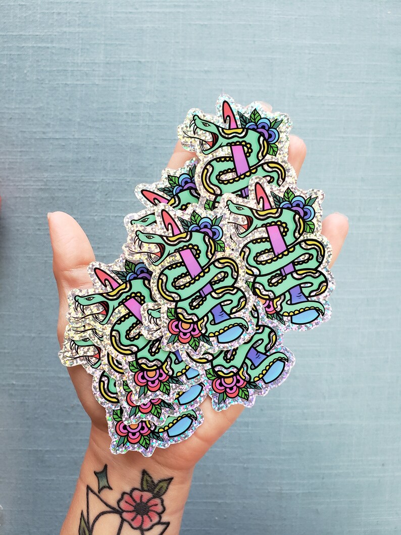 May include: A handful of holographic stickers featuring a green snake with a pink dagger through its body, surrounded by flowers and leaves. The stickers have a glittery, iridescent finish and are on a light blue background.