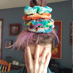 May include: A stack of four colorful crocheted hair scrunchies in shades of blue, green, orange, and pink. The scrunchies are stacked on top of each other and are being worn by a person with pink hair.