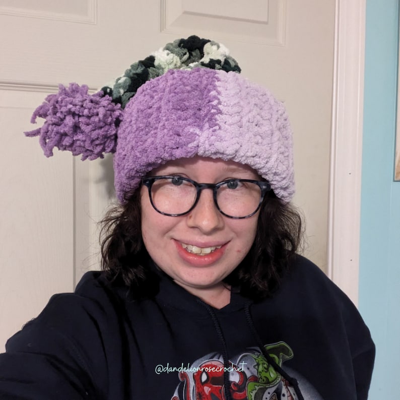 Op de afbeelding: Een persoon draagt ​​een paarse en groene gebreide muts met een pompon. De muts is gemaakt van pluche garen en heeft een uniek ontwerp. De persoon draagt ​​een zwarte hoodie met een afbeelding op de voorkant. De tekst op de hoodie luidt "@dandelionrosecrochet".