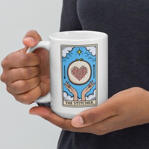 May include: White ceramic mug with a blue and white illustration of a hand holding a hoop with a red heart cross-stitched inside. The text "THE STITCHER." is printed below the illustration.