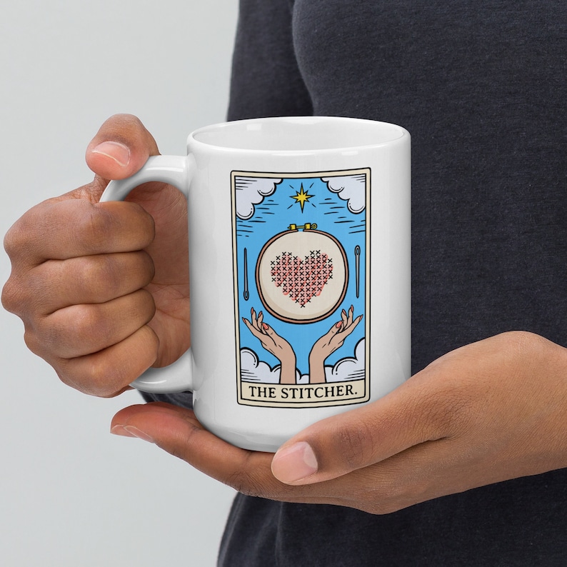 May include: White ceramic mug with a blue and white illustration of a hand holding a hoop with a red heart cross-stitched inside. The text "THE STITCHER." is printed below the illustration.