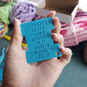 Op de afbeelding: Een turquoise garenmeter wordt getoond voor een verzameling kleurrijke bollen wol en breiaccessoires. De meter toont garendiktes: Super Bulky 5-6, Bulky 7, Aran 8, Worsted 9, DK 11, Sport 12, Fingering 14+ en Lace 30+.