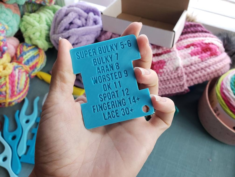 Op de afbeelding: Een turquoise garenmeter wordt getoond voor een verzameling kleurrijke bollen wol en breiaccessoires. De meter toont garendiktes: Super Bulky 5-6, Bulky 7, Aran 8, Worsted 9, DK 11, Sport 12, Fingering 14+ en Lace 30+.